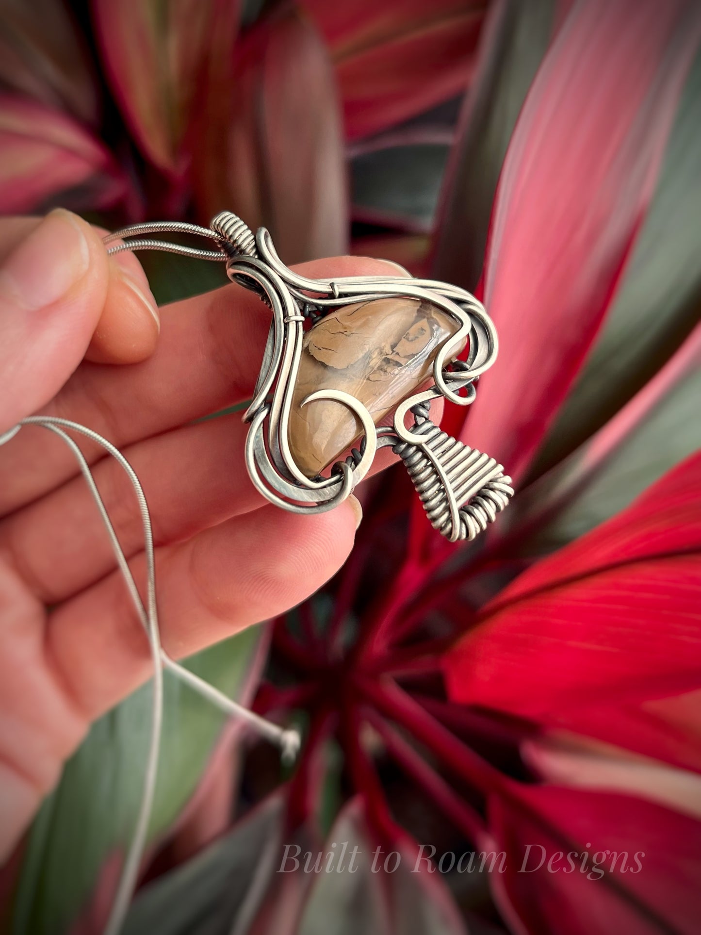 Hand holding a silver mushroom-shaped pendant with a brown Mookaite Jasper gemstone with plant background - side view