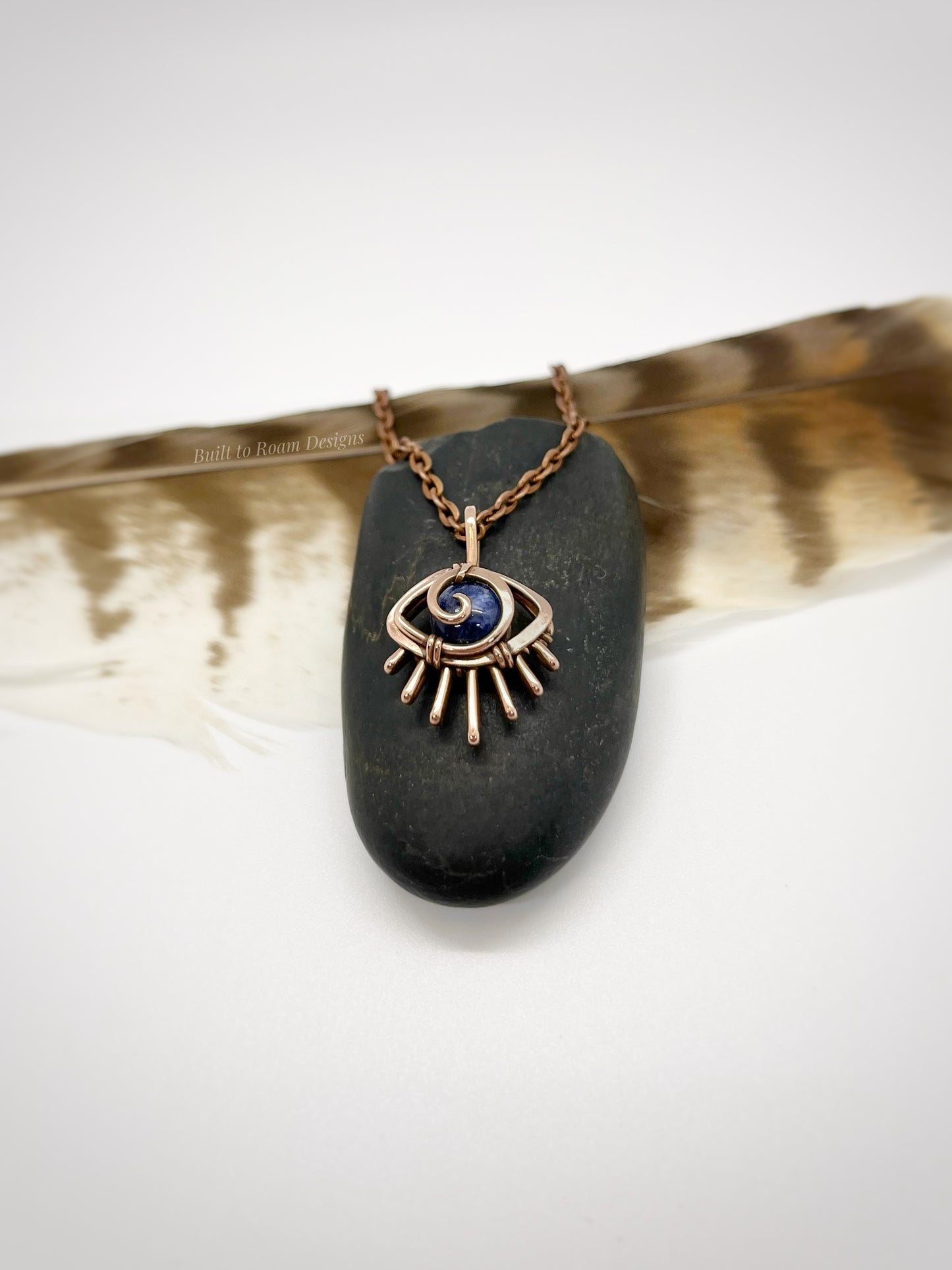 Copper eye-shaped pendant necklace with a blue Sodalite gemstone on a black stone, placed on a feather - side view - Insight