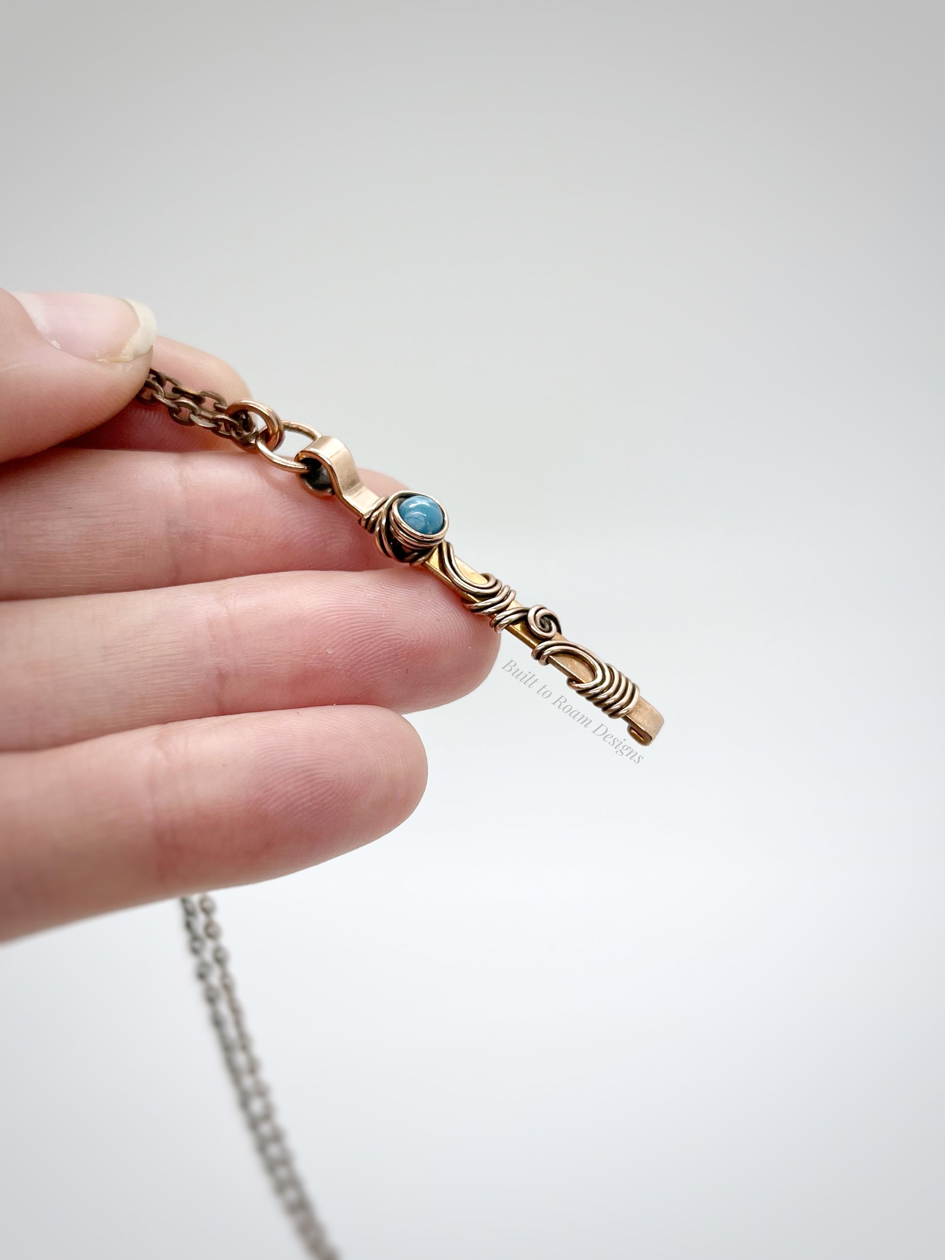 Hand holding a swirly copper bar necklace with a blue Larimar bead with white background - side view - Current