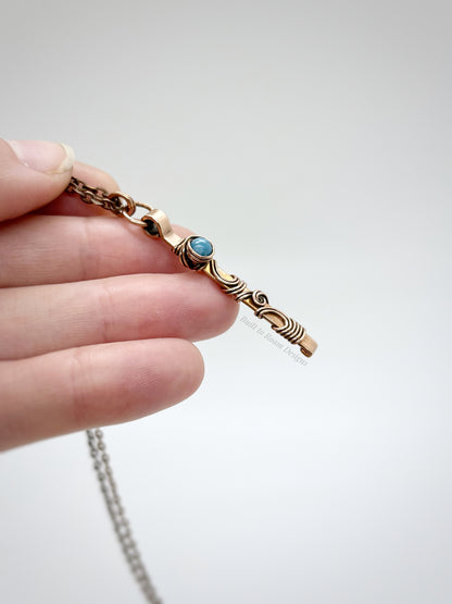 Hand holding a swirly copper bar necklace with a blue Larimar bead with white background - side view - Current