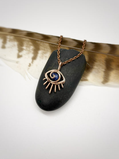 Copper eye-shaped pendant necklace with a blue Sodalite gemstone on a black stone, placed on a feather - side view - Insight