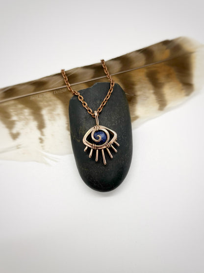 Copper eye-shaped pendant necklace with a blue Sodalite gemstone on a black stone, placed on a feather - front view - Insight