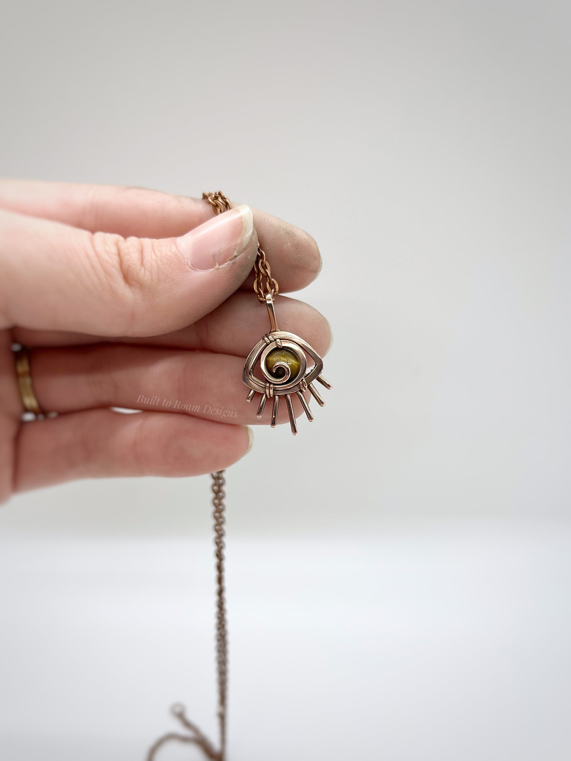 Hand holding a Copper eye-shaped pendant necklace with a golden Tiger’s Eye gemstone against a white background - side view - Guardian