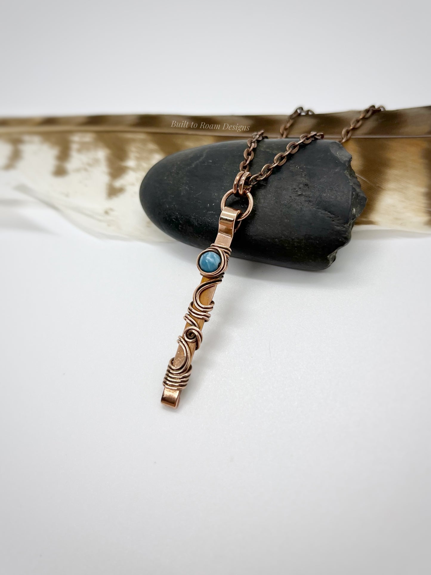 Swirly copper bar necklace with a blue Larimar bead on a rock with white background - side view - Current