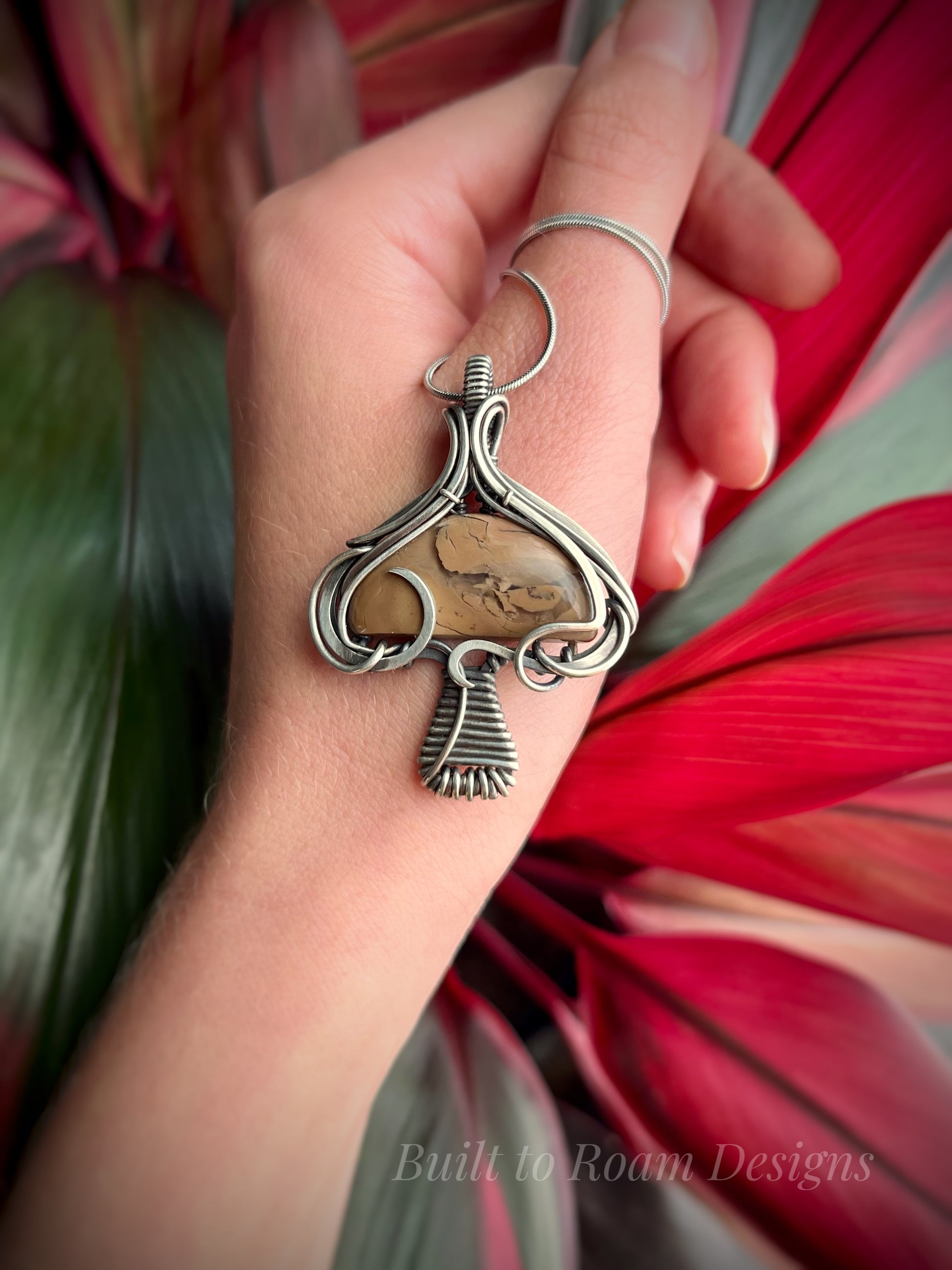 Hand holding a silver mushroom-shaped pendant with a brown Mookaite Jasper gemstone with plant background - front view