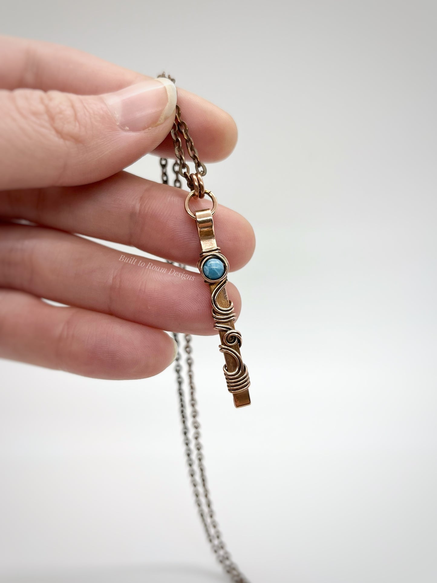Hand holding a swirly copper bar necklace with a blue Larimar bead with white background - front view - Current