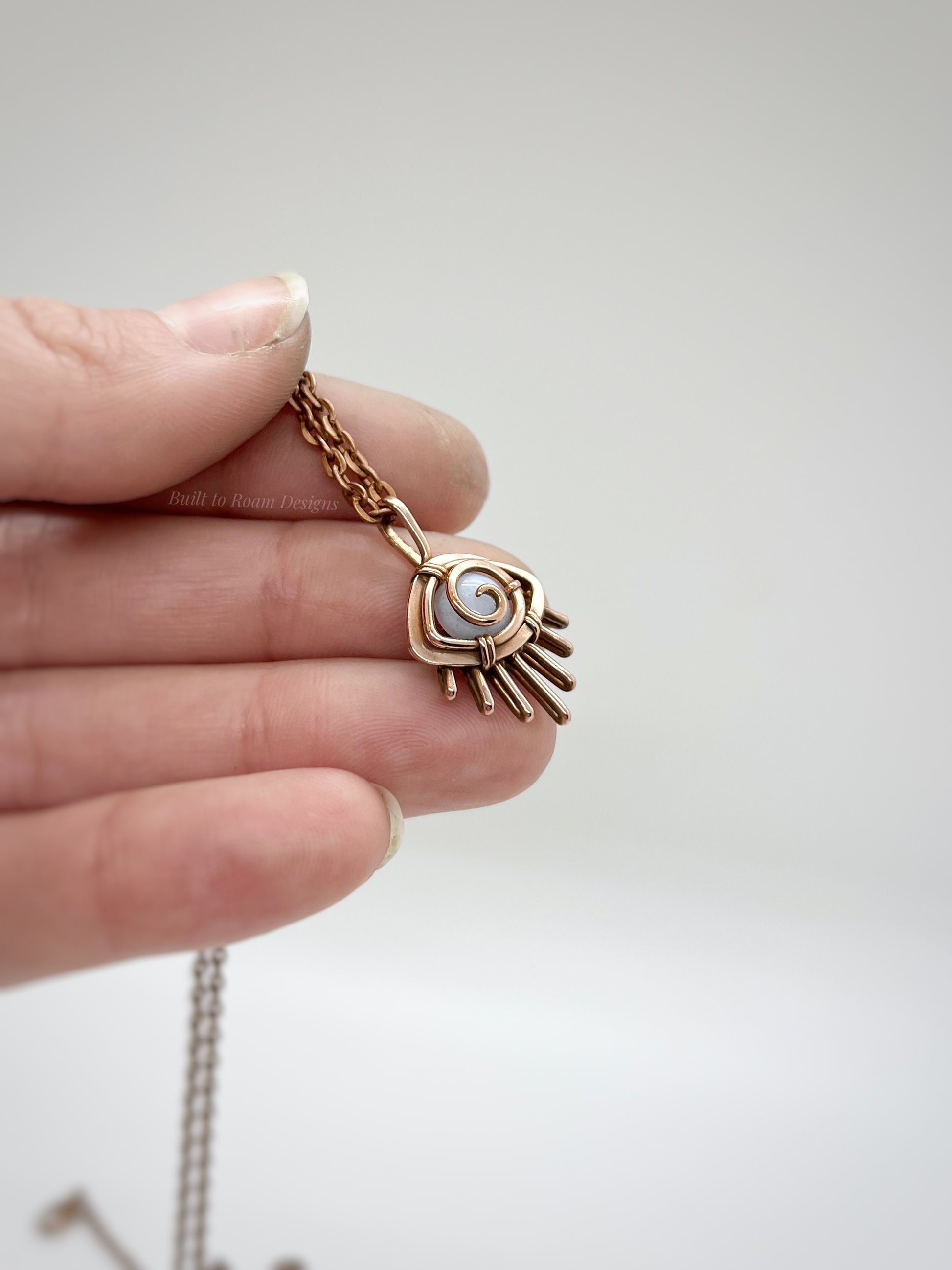 Hand holding a Copper eye-shaped pendant necklace with a blue lace agate gemstone against a white background - side view - Comfort