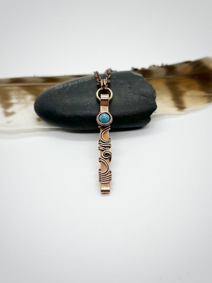 Swirly copper bar necklace with a blue Larimar bead on a rock with white background - side view - Current