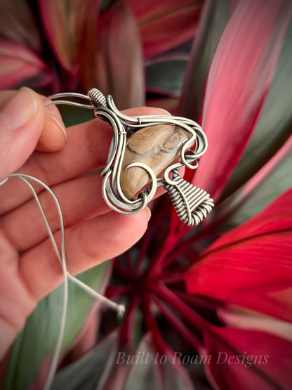 Hand holding a silver mushroom-shaped pendant with a brown Mookaite Jasper gemstone with plant background - side view