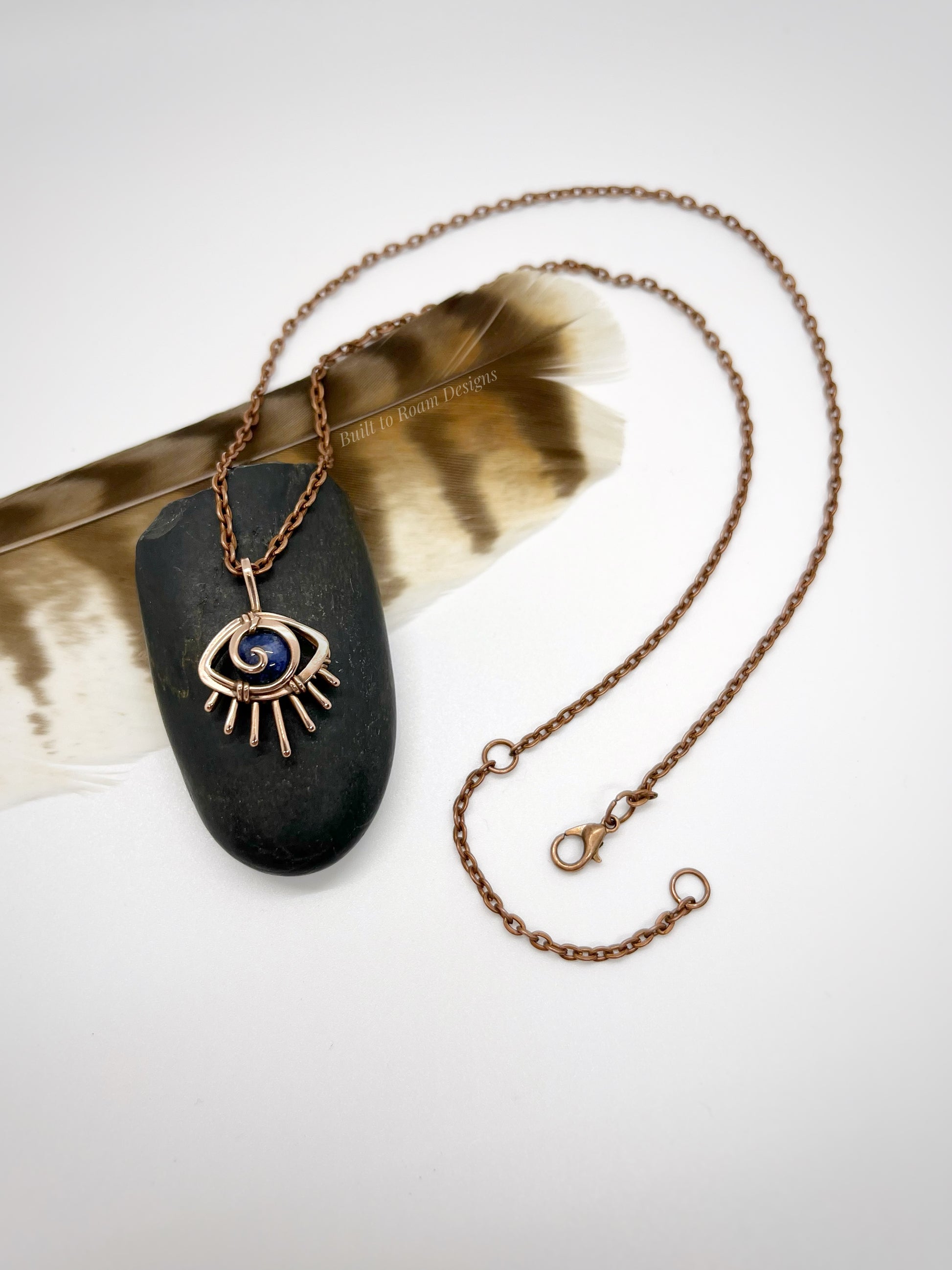 Copper eye-shaped pendant necklace with a blue Sodalite gemstone on a black stone, placed on a feather - side view - Insight