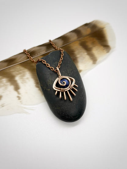 Copper eye-shaped pendant necklace with a blue Sodalite gemstone on a black stone, placed on a feather - side view - Insight