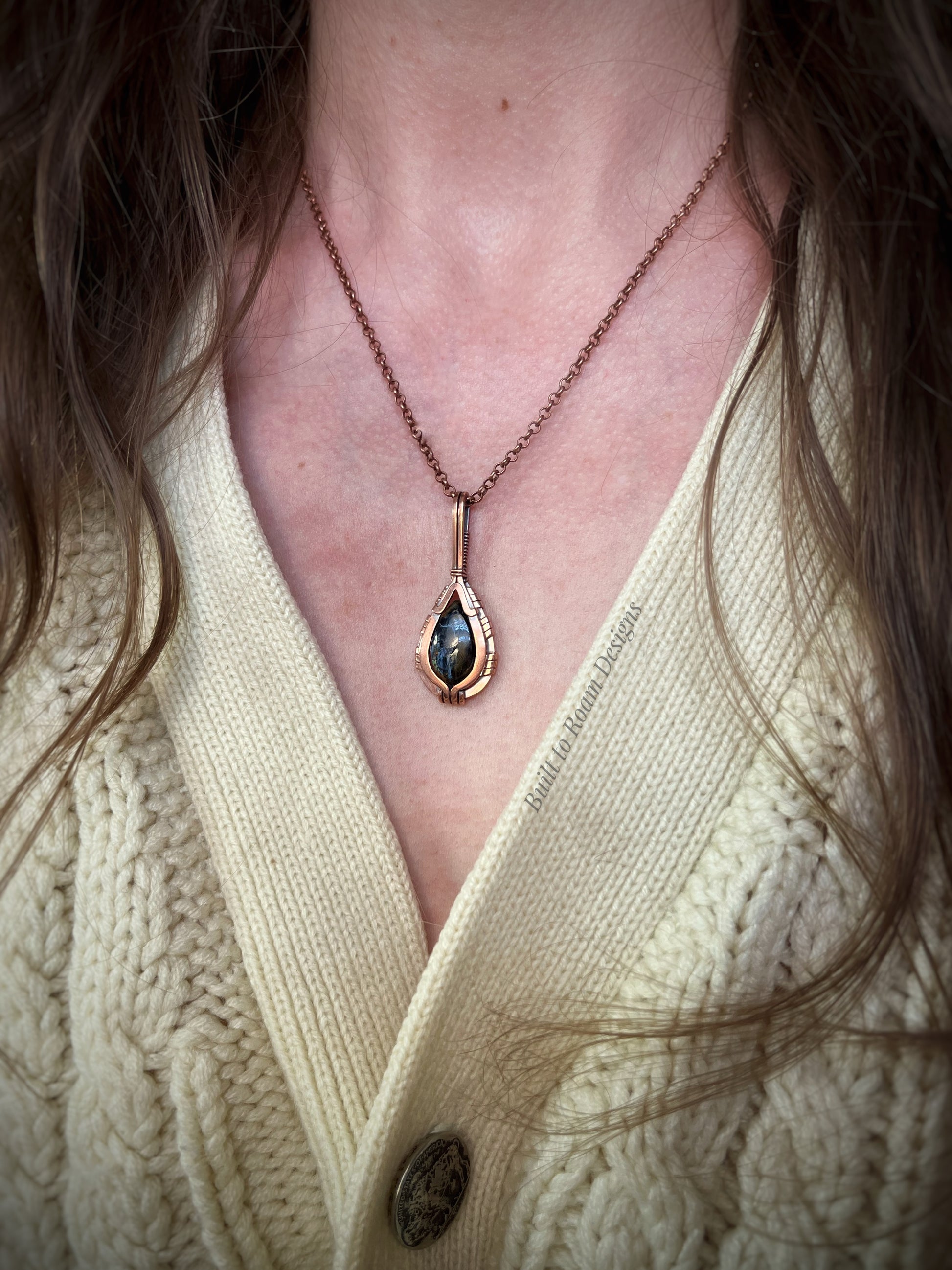 Pietersite in copper teardrop pendant worn by a person in a light-colored sweater.