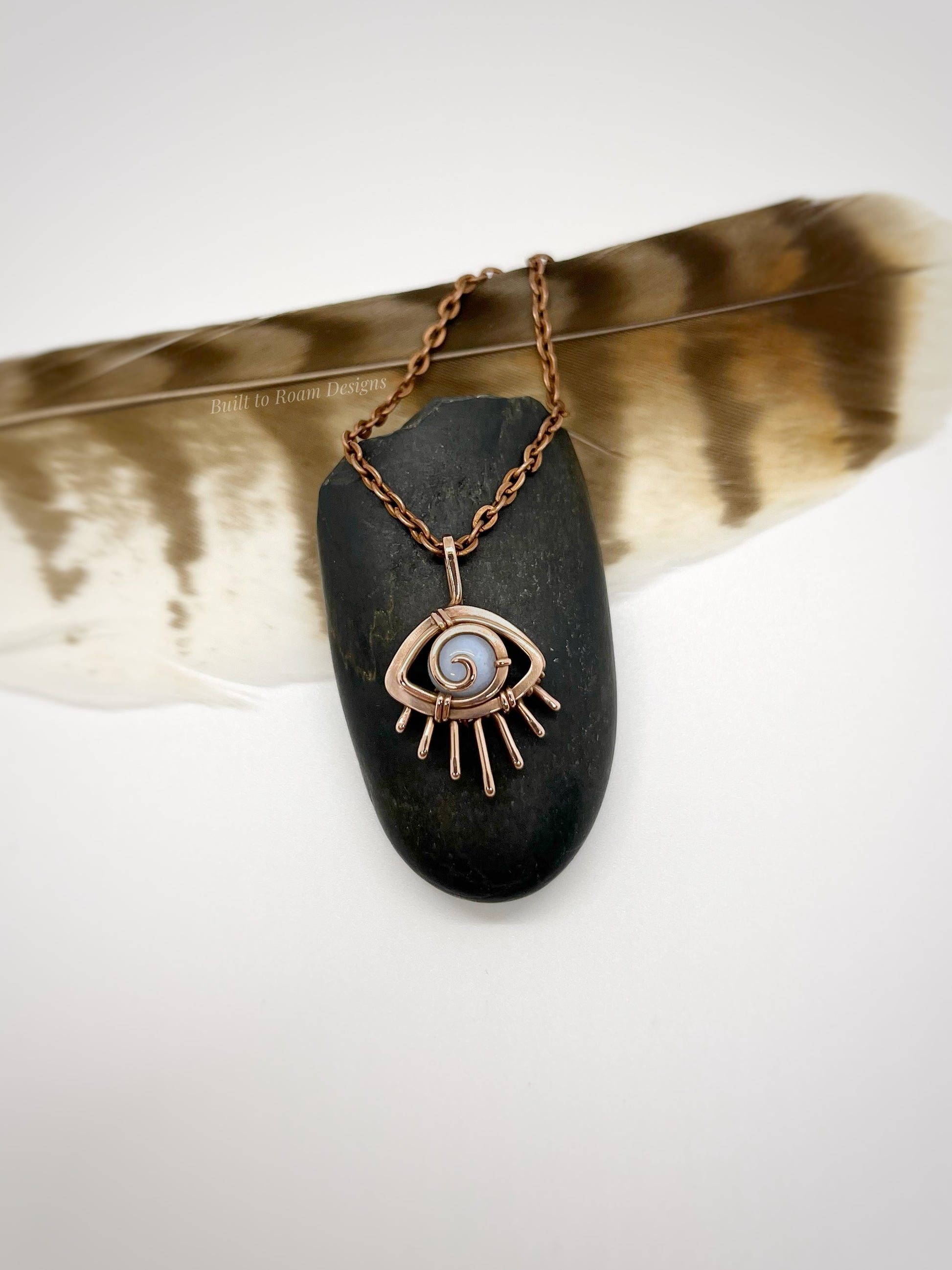 Copper eye-shaped pendant necklace with a blue lace agate gemstone against a dark stone with white background - front view - Comfort