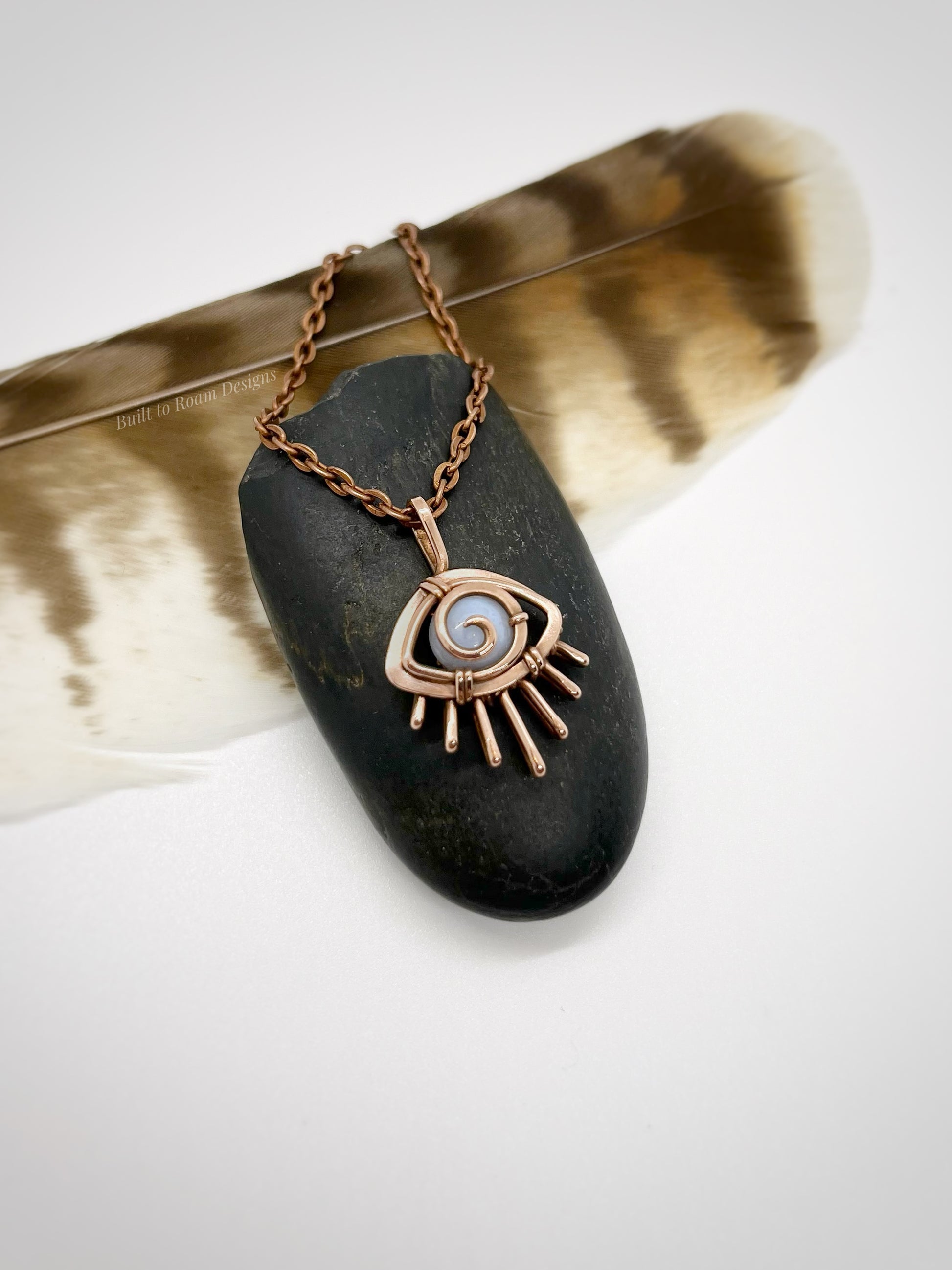 Copper eye-shaped pendant necklace with a blue lace agate gemstone against a dark stone with white background - side view - Comfort