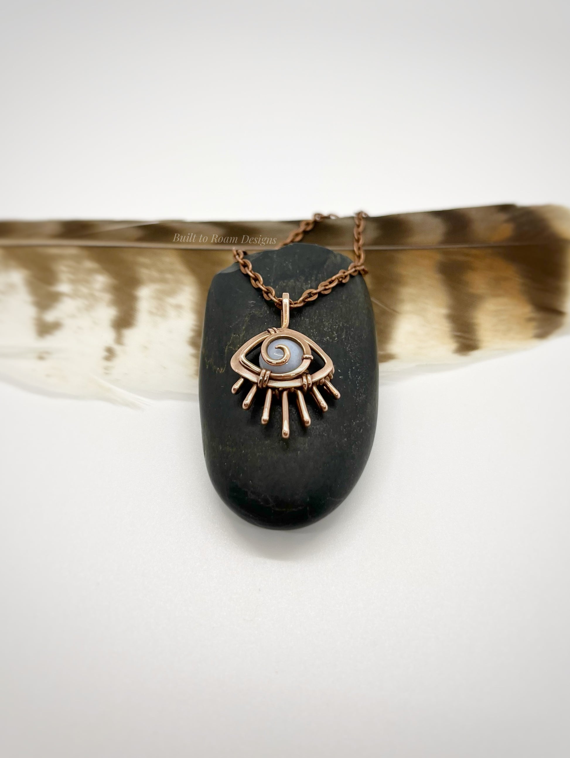 Copper eye-shaped pendant necklace with a blue lace agate gemstone against a dark stone with white background - side view - Comfort