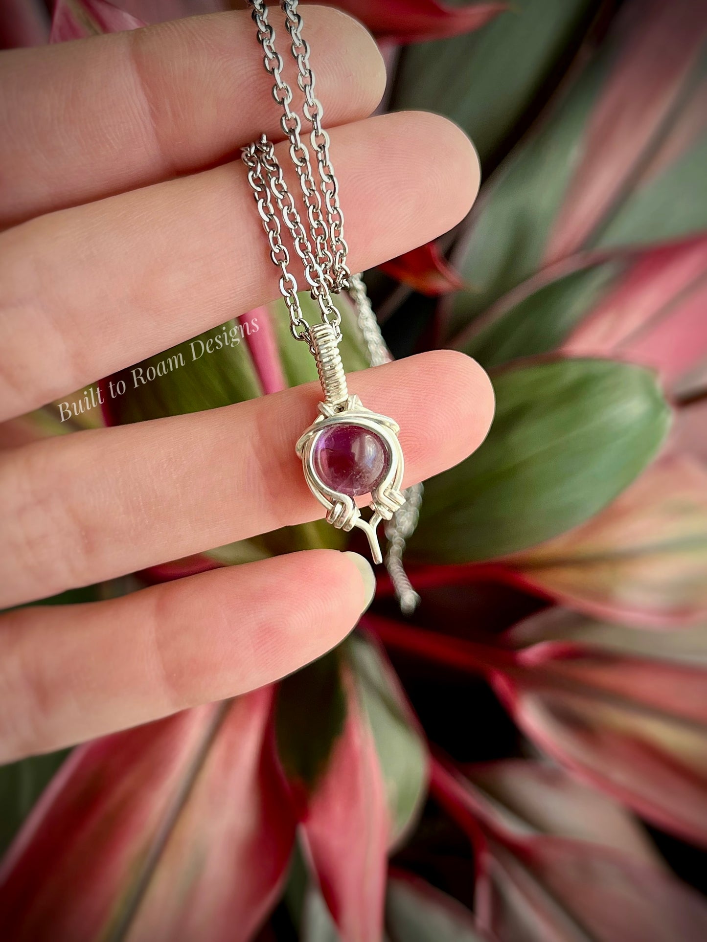 Silver necklace with a purple amethyst gemstone held between fingers against a plant background