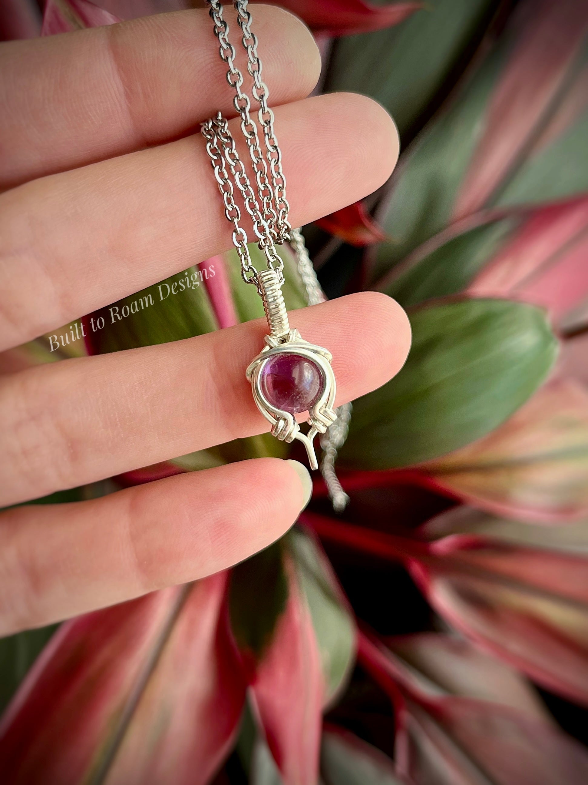 Silver necklace with a purple amethyst gemstone held between fingers against a plant background