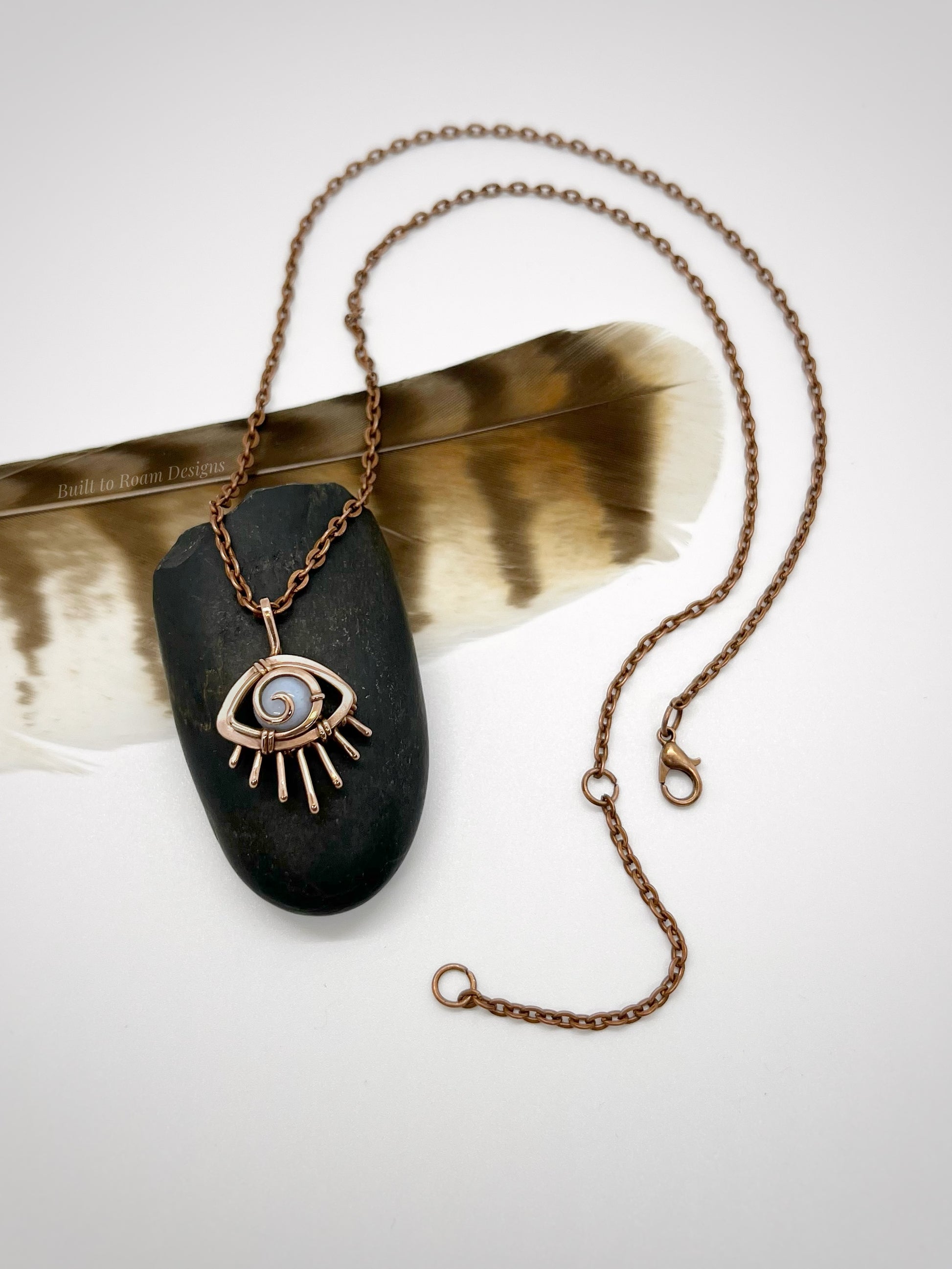 Copper eye-shaped pendant necklace with a blue lace agate gemstone against a dark stone with white background - front view - Comfort