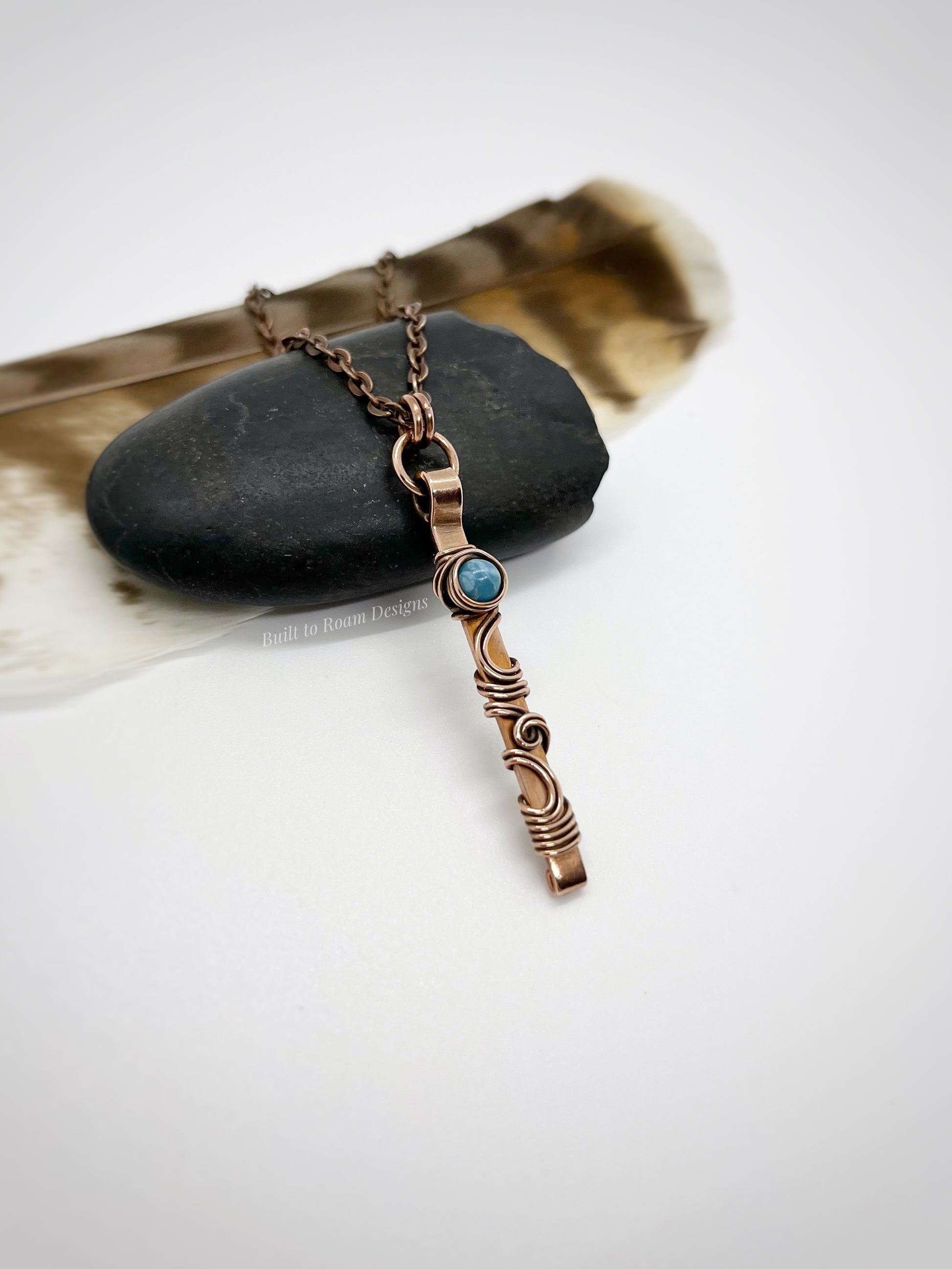 Swirly copper bar necklace with a blue Larimar bead on a rock with white background - side view - Current