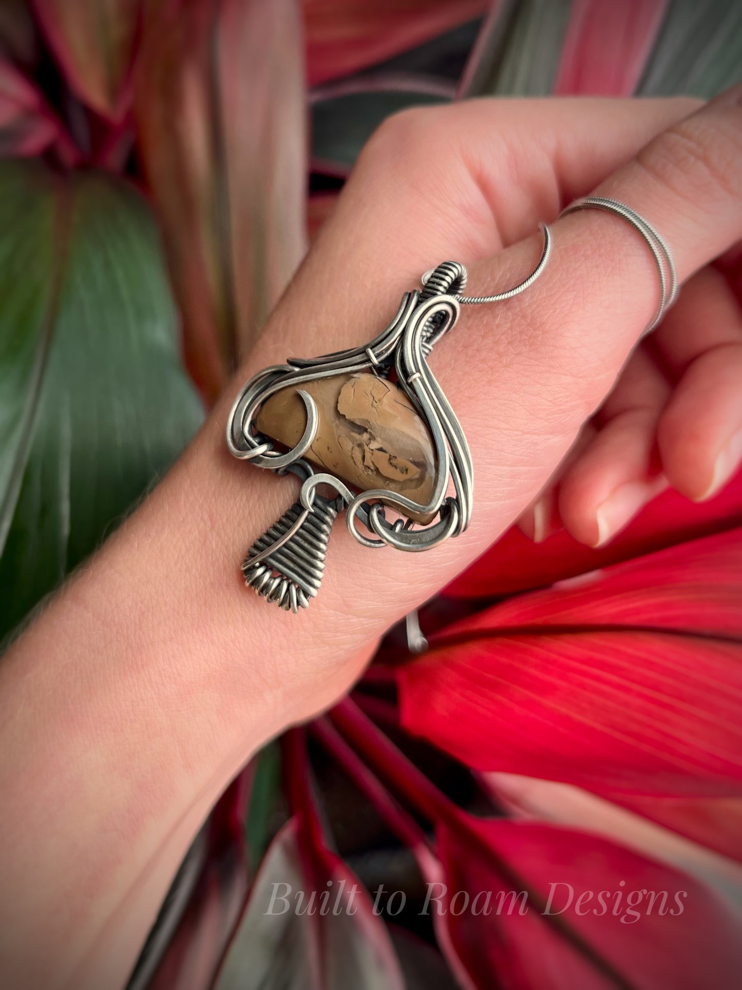 Hand holding a silver mushroom-shaped pendant with a brown Mookaite Jasper gemstone with plant background - side view