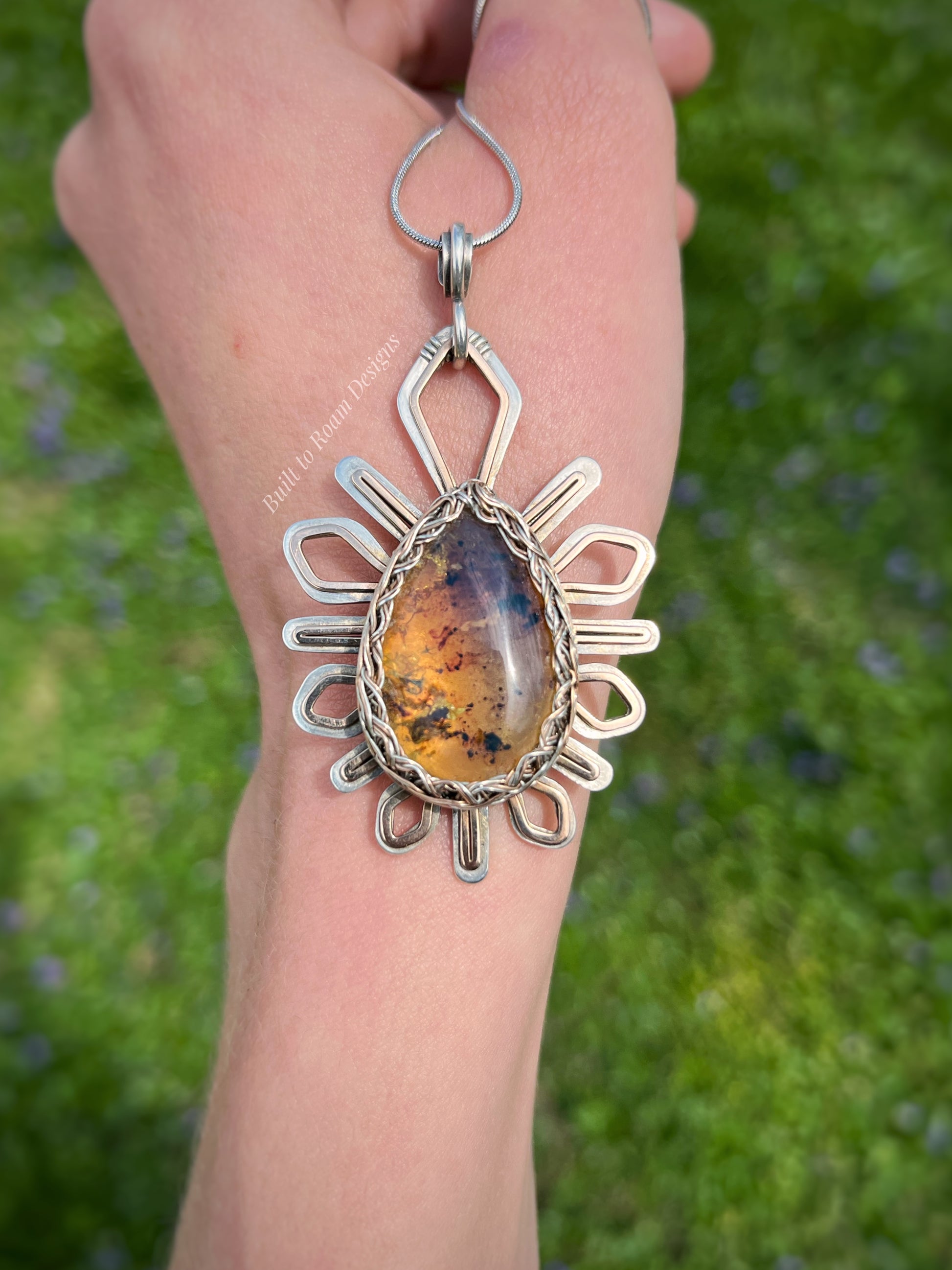 Front view of hand holding a silver and bronze star-burst pendant with a golden amber gemstone - Liora