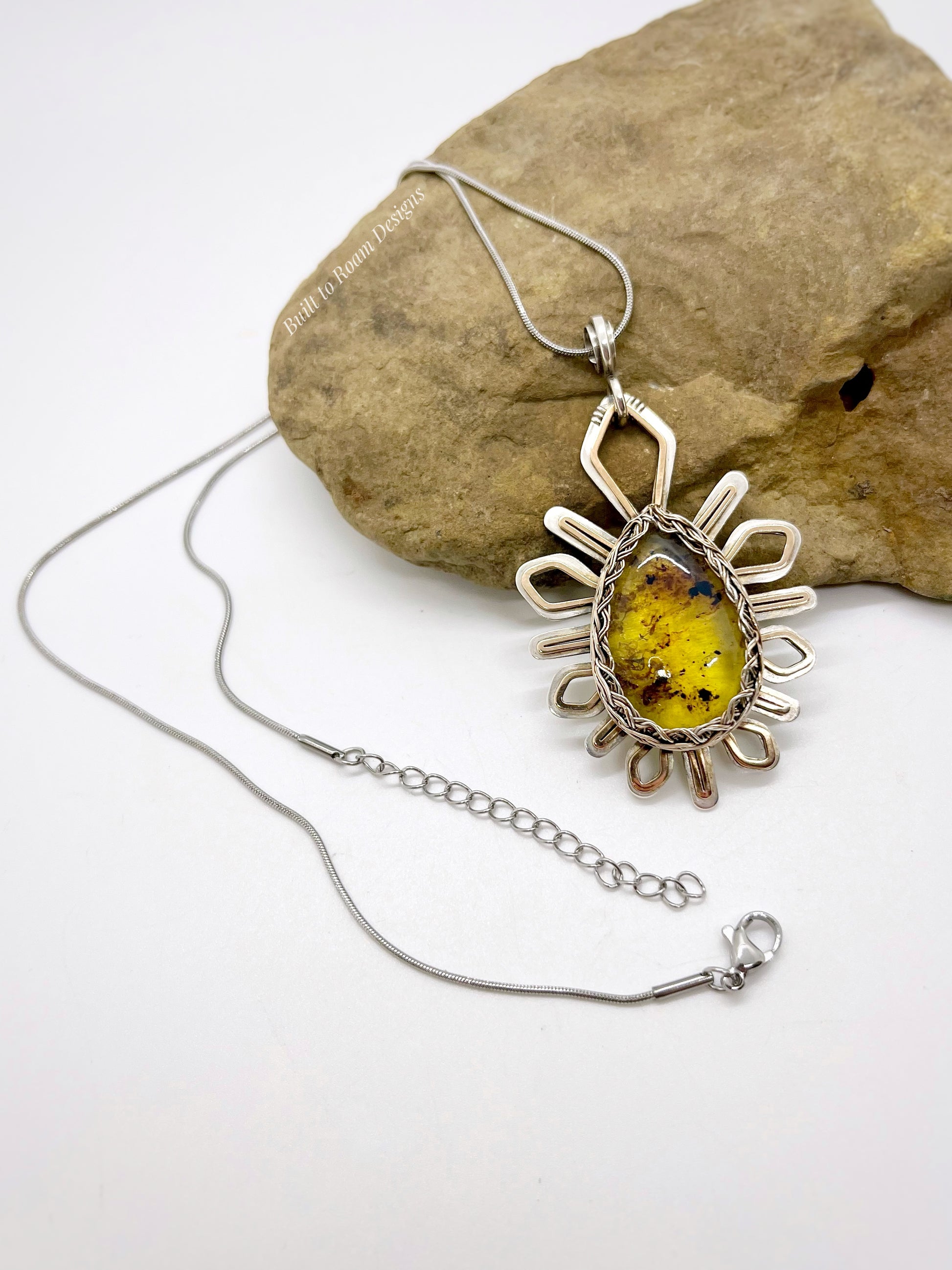 Front view on a rock of a silver and bronze star-burst pendant with a golden amber gemstone on a necklace - Liora