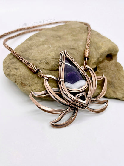 Side view of a copper lotus flower Viking knit chain necklace with purple amethyst gemstone on a rock - Elysian