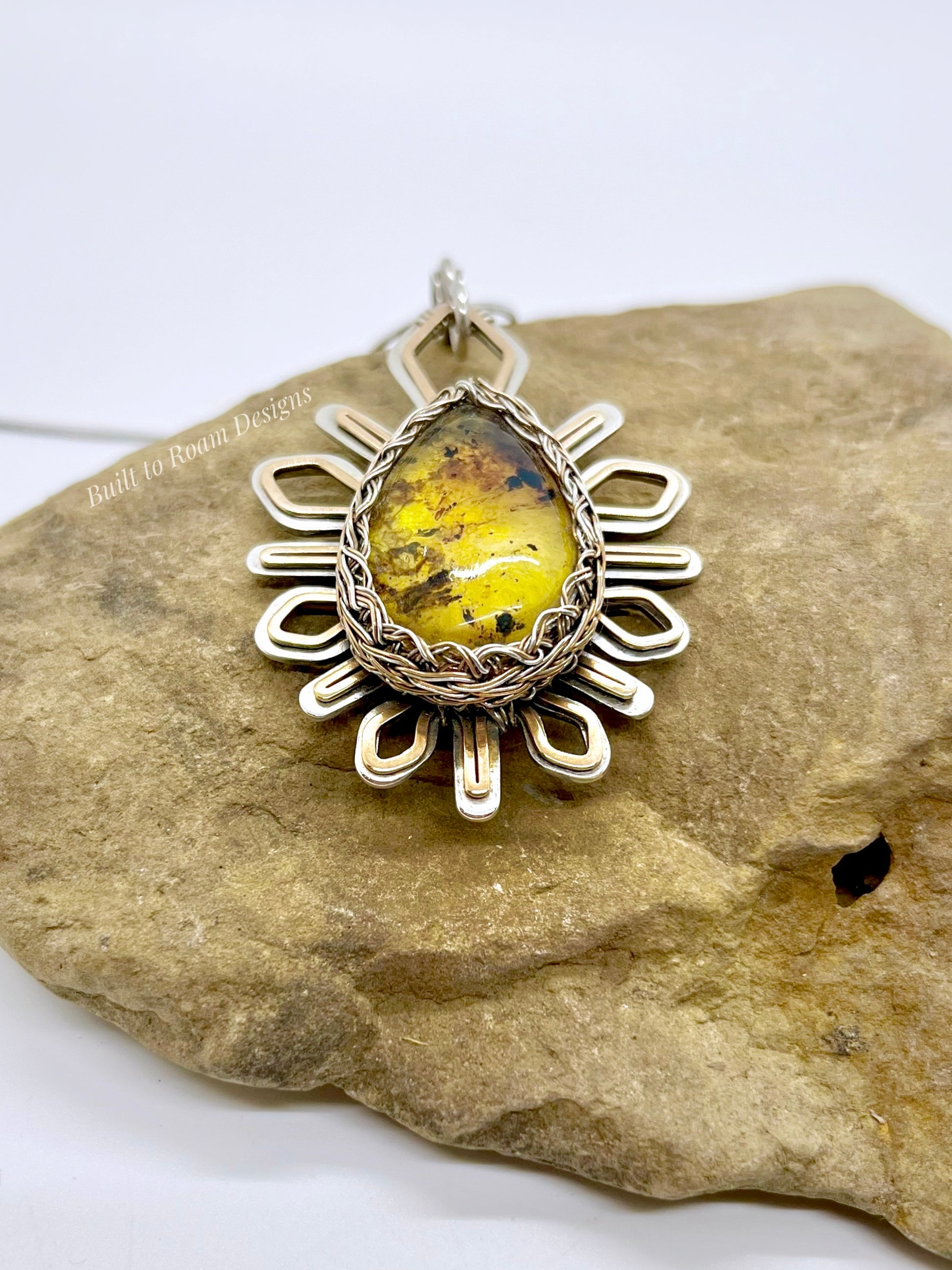 Silver and bronze star-burst pendant with a golden amber gemstone on a rock - Liora