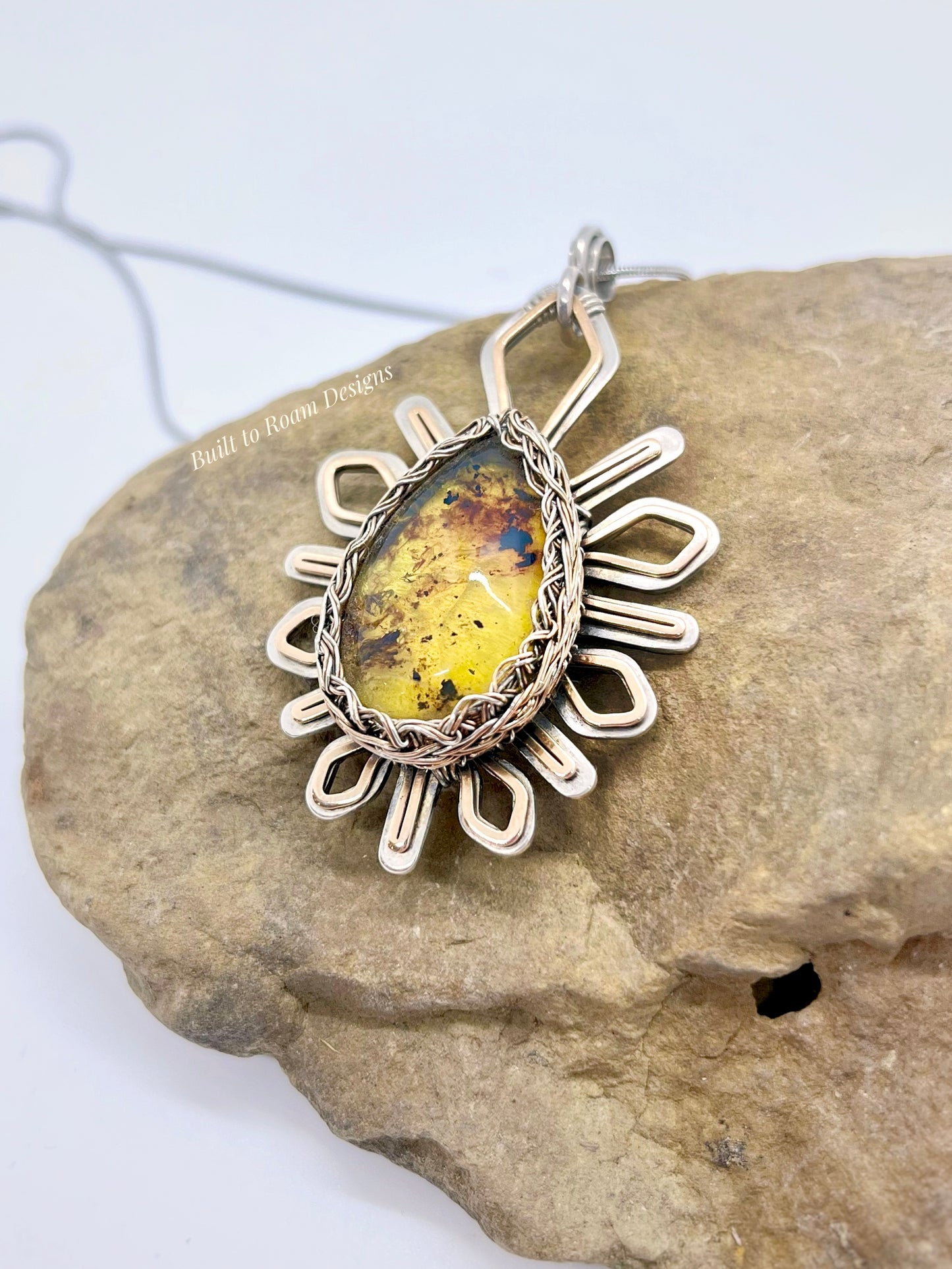 Silver and bronze star-burst pendant with a golden amber gemstone on a rock - Liora