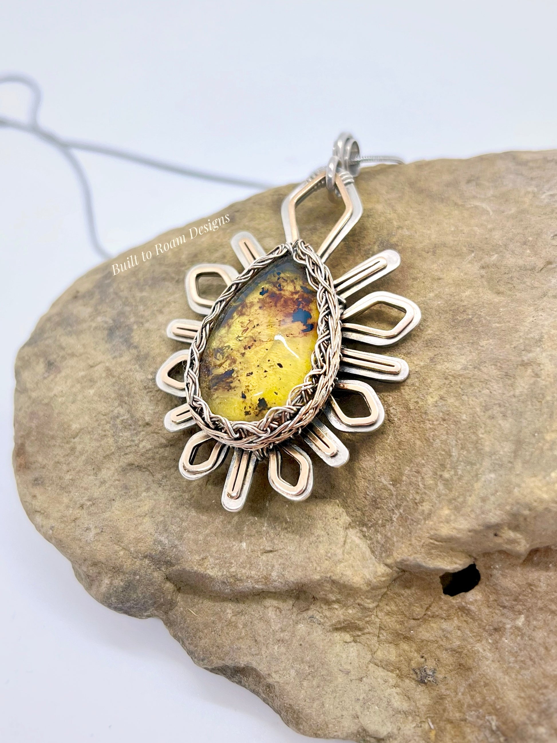 Silver and bronze star-burst pendant with a golden amber gemstone on a rock - Liora