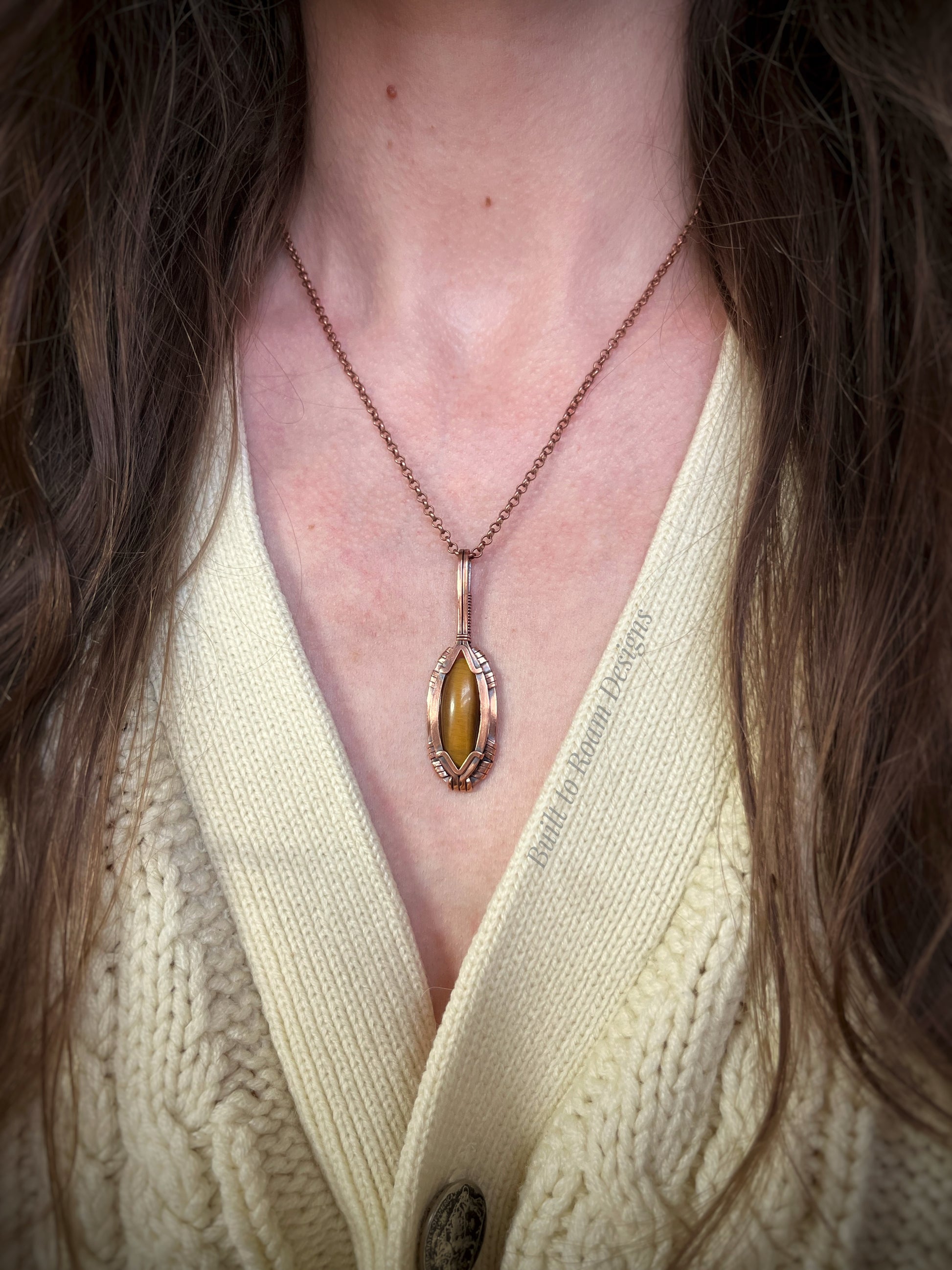 Necklace with a tiger's eye pendant in copper worn by a person in a light-colored sweater.
