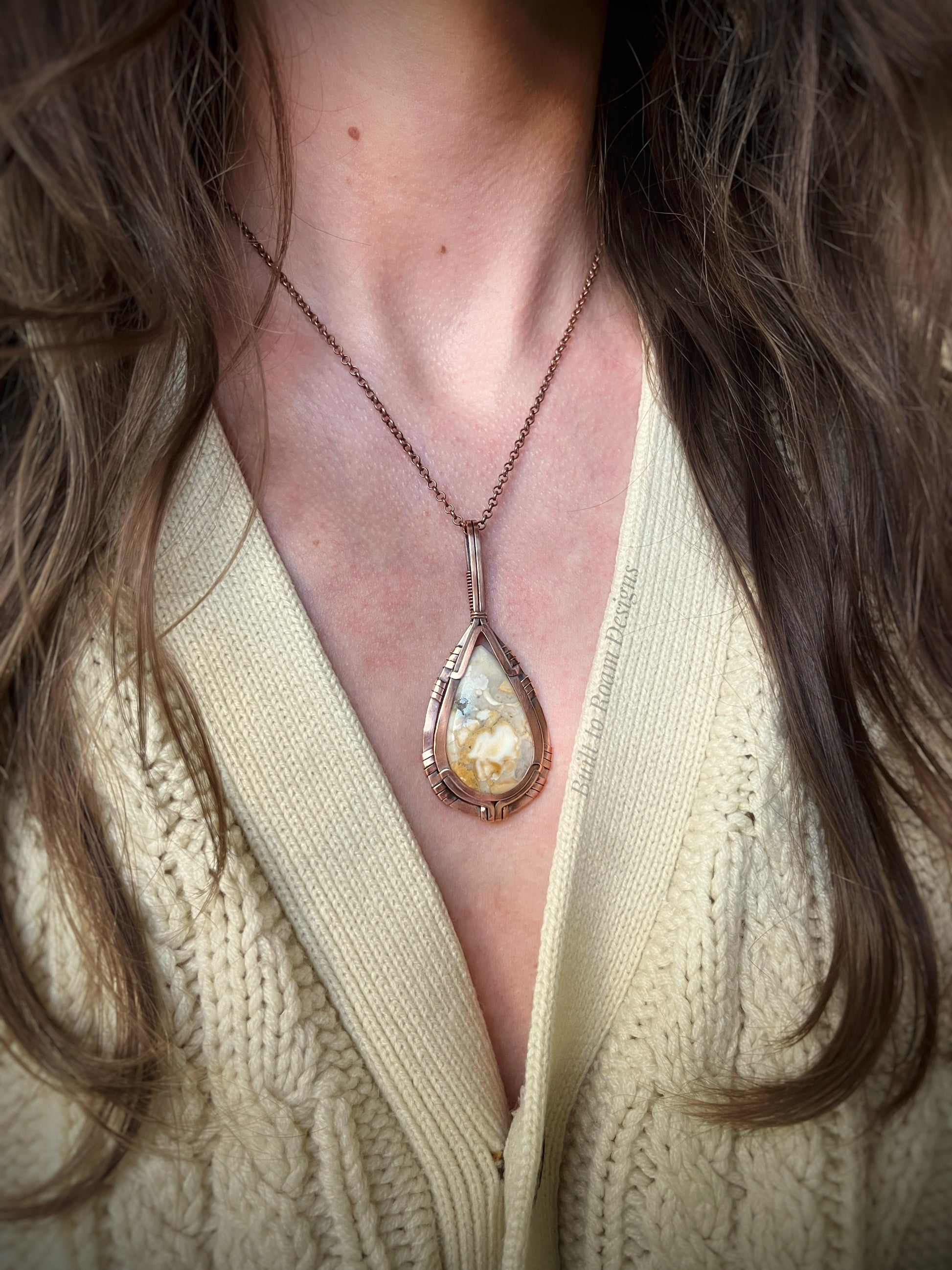 Close-up of a necklace with a teardrop-shaped wild horse jasper pendant in copper worn by a person in a beige sweater.