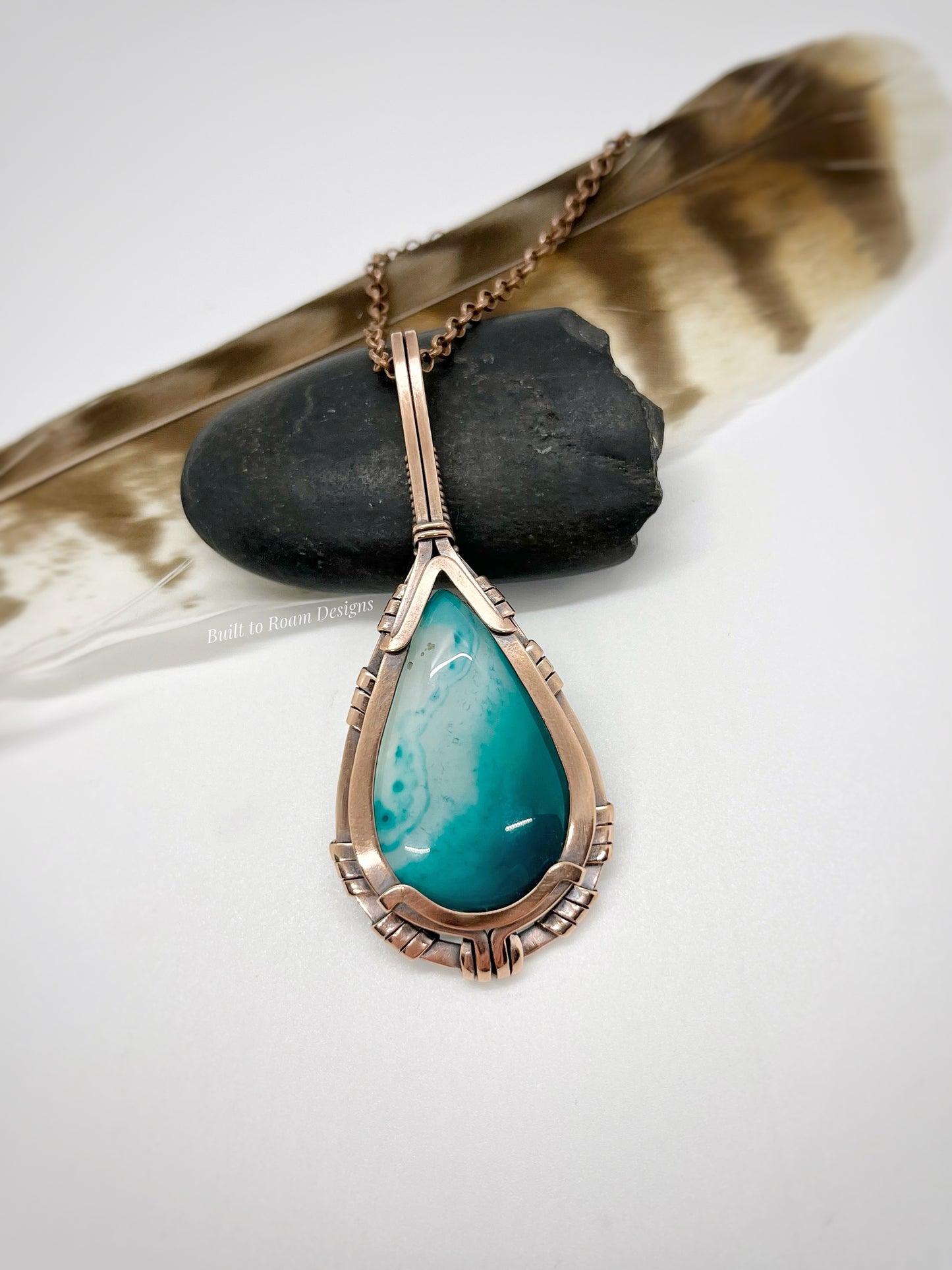 Teardrop-shaped turquoise color enhanced banded agate pendant in copper on a chain with a stone and feather in the background.